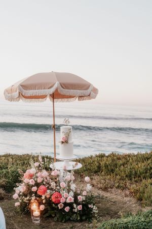 Beach wedding setup with vintage umbrella, pink roses, and ocean view at sunset.