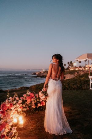 Sunset wedding scene with bride standing among illuminated coastal flowers.