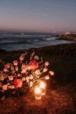 Pink roses and glowing candles arranged on a cliff overlooking the ocean at dusk.