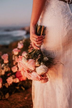 A white wedding dress and pink bouquet captured at sunset against an ocean backdrop.