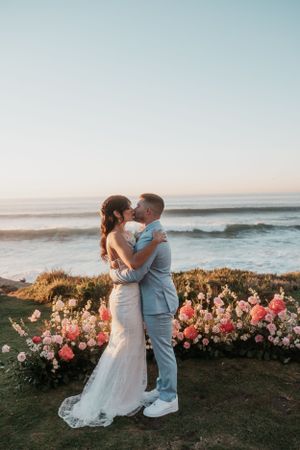 Newlyweds embrace by the ocean with pink floral arrangements during sunset.