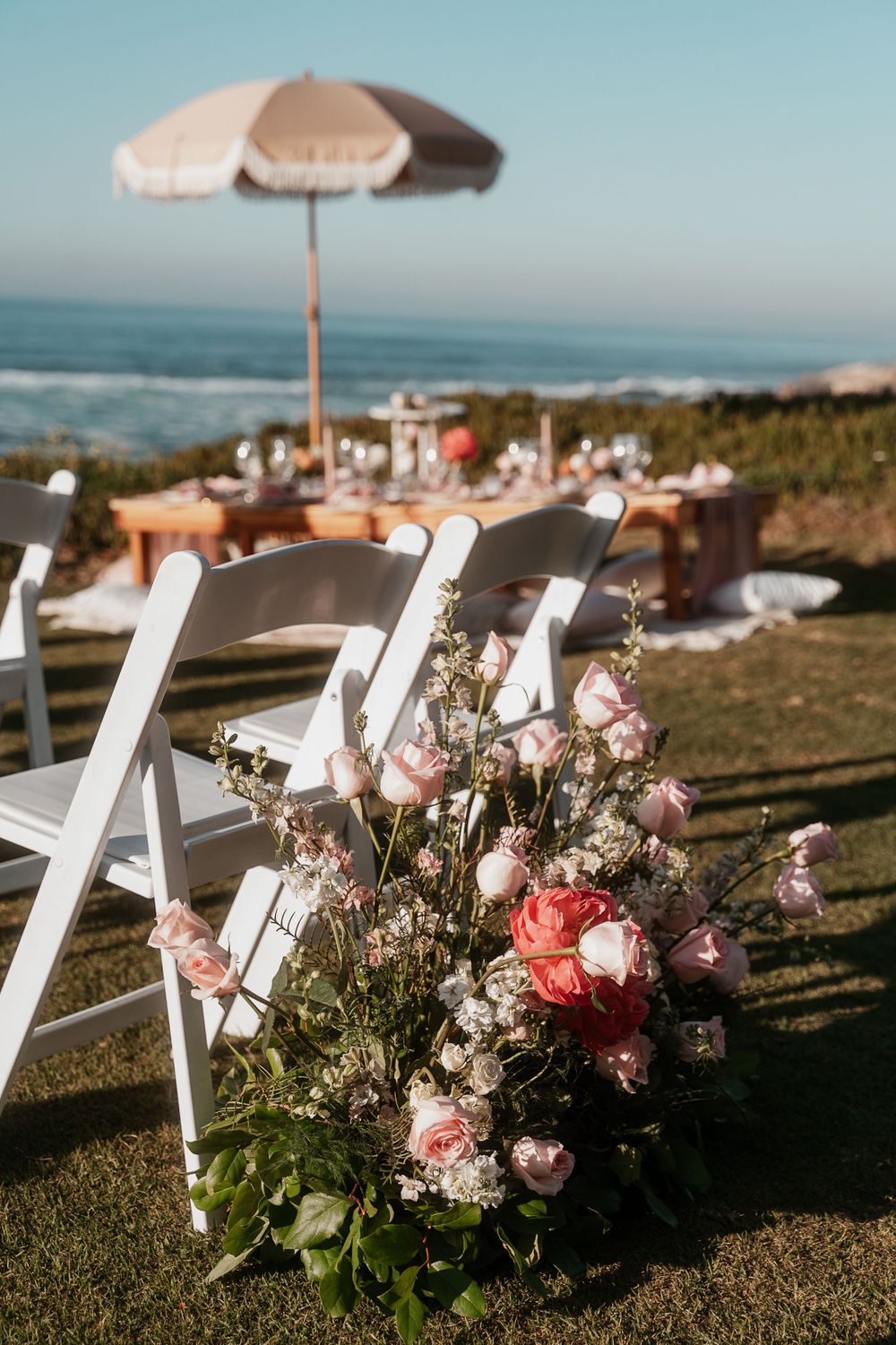 Oceanside wedding ceremony setup with white chairs and pink floral aisle decorations.