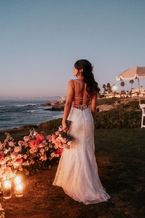 Bride in backless lace gown stands among flowers at oceanside sunset.
