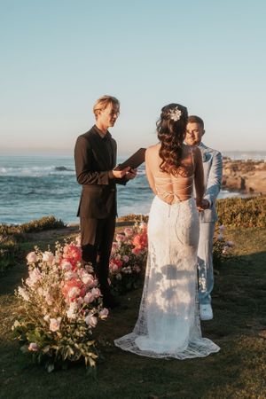 An officiant conducts a wedding ceremony on a coastal cliff with pink floral arrangements.