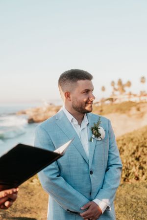 A groom in a light blue suit stands during an outdoor coastal wedding ceremony at sunset.