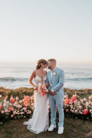 Romantic beach wedding ceremony setup with pink and white floral arrangements against an ocean backdrop at sunset.