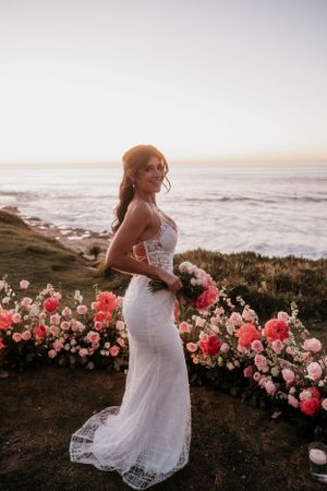 Bride poses in lace wedding dress among pink roses by oceanside cliff.