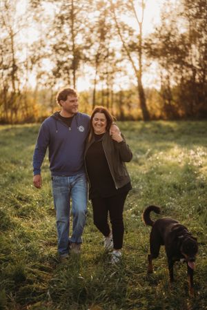 A couple walks their black dog through a scenic field during sunset.