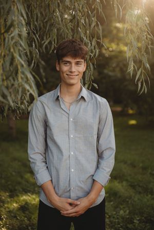 Outdoor portrait of young person in grey button-down shirt standing in front of willow tree at golden hour.