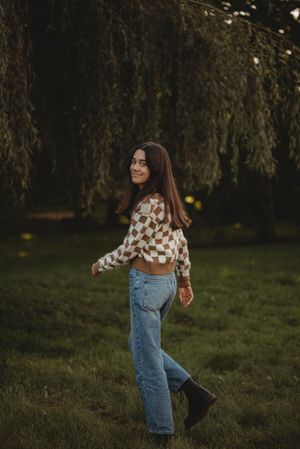 A person in jeans and a patterned sweater poses under a willow tree at dusk.