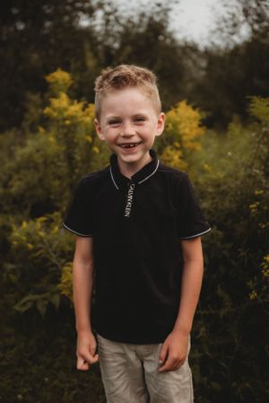Young child in black polo shirt smiles while standing outdoors with yellow flowers in background.