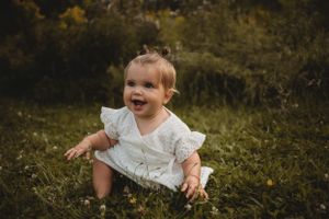 Joyful baby in white dress sitting on grass during outdoor portrait session.