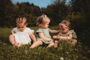 Three babies sitting together in grass wearing summer outfits.
