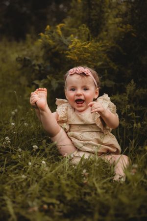 Baby in cream lace dress sits joyfully in grass against blurred yellow flowers in summer garden.