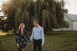 A couple holds hands while walking in a backyard with willow tree backdrop at sunset.