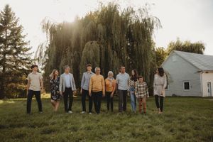 A line of people stands in front of a weeping willow tree and white house during sunset.