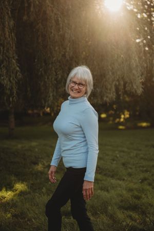Senior person in light blue turtleneck sweater standing outdoors at sunset with willow trees.