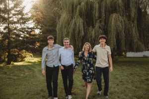 Four people stand together outdoors with a weeping willow tree and white building in background.