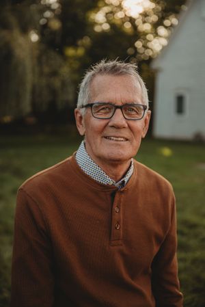 Senior person wearing glasses and brown sweater smiling outdoors with white building in background.