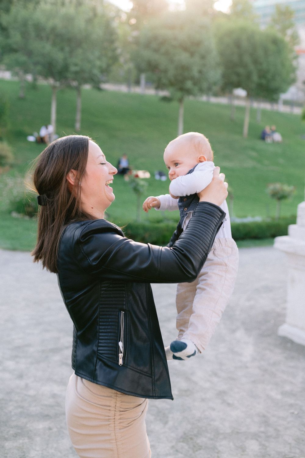 A joyful mother in a black leather jacket lifts her smiling baby in a sunny green park.