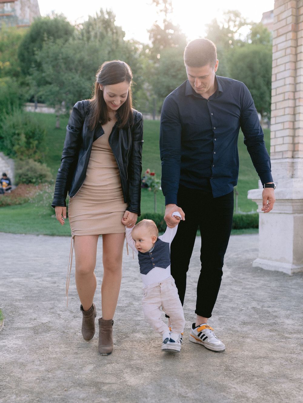 A young family with a toddler walks hand-in-hand on a stone path in a sunlit park.