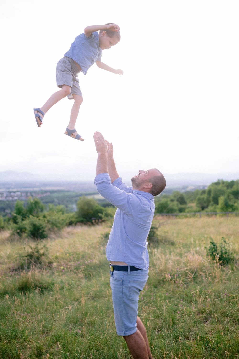 Father joyfully tossing young child high in the air in a green meadow with scenic landscape behind.