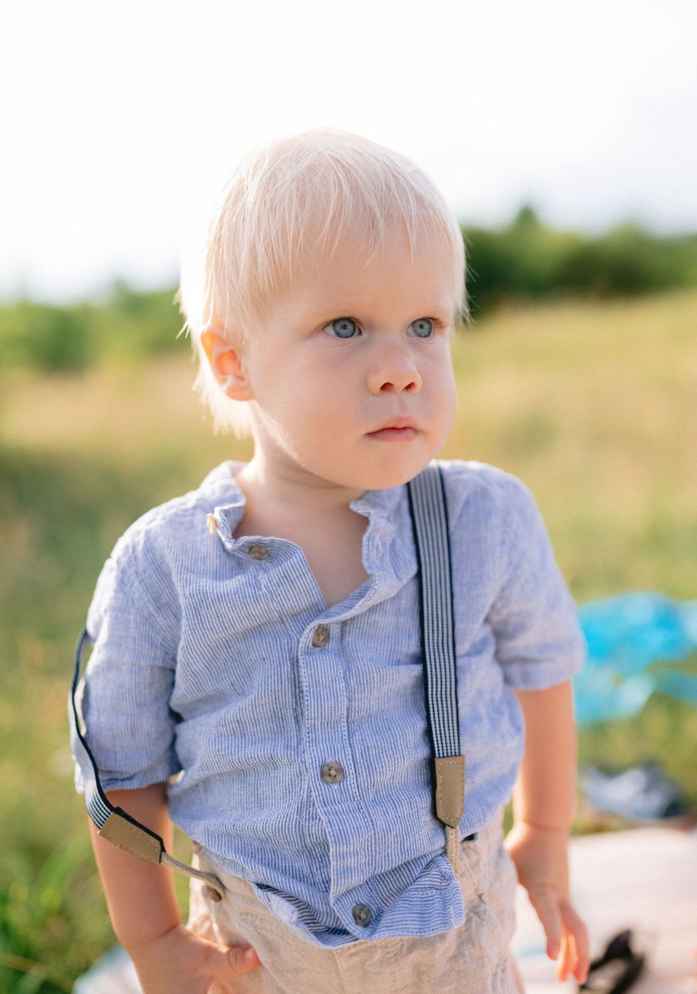 Adorable blonde toddler boy in blue linen shirt with suspenders standing outdoors in soft sunlight.