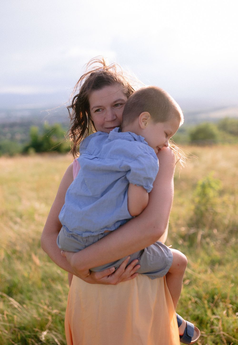 Mother tenderly holding and hugging her young child in a golden sunlit meadow with hills in background.