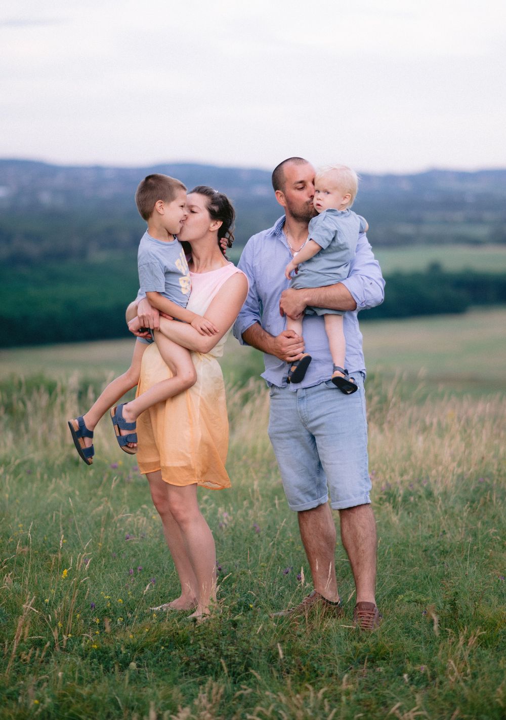 Happy family of four with two young children sharing kisses in a lush green meadow at dusk.