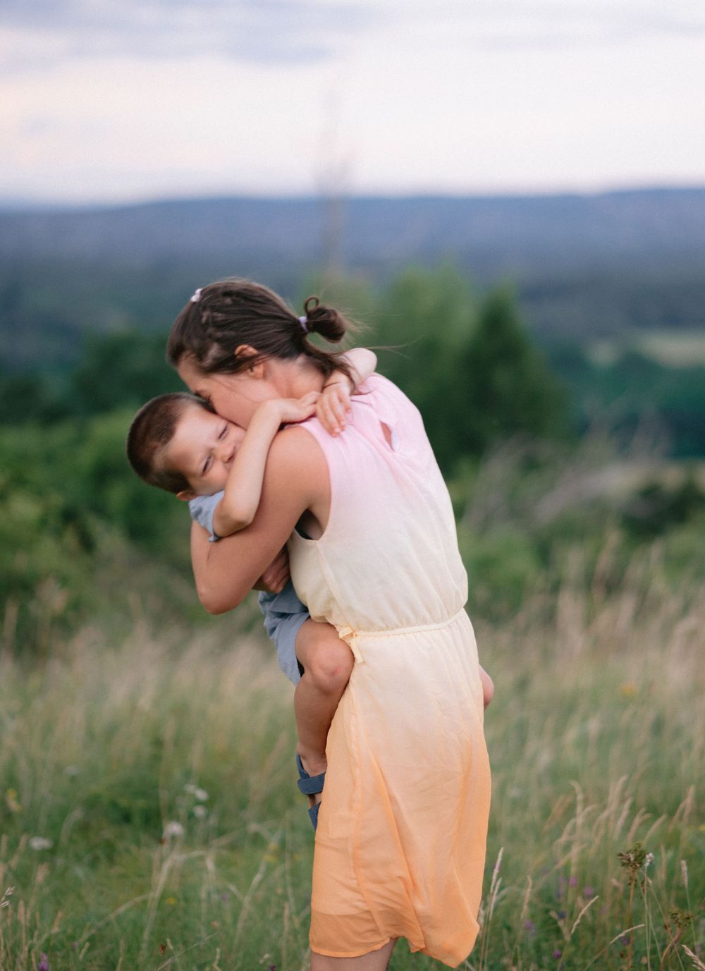 A young girl lovingly hugs her little brother while standing in a lush green field at dusk.