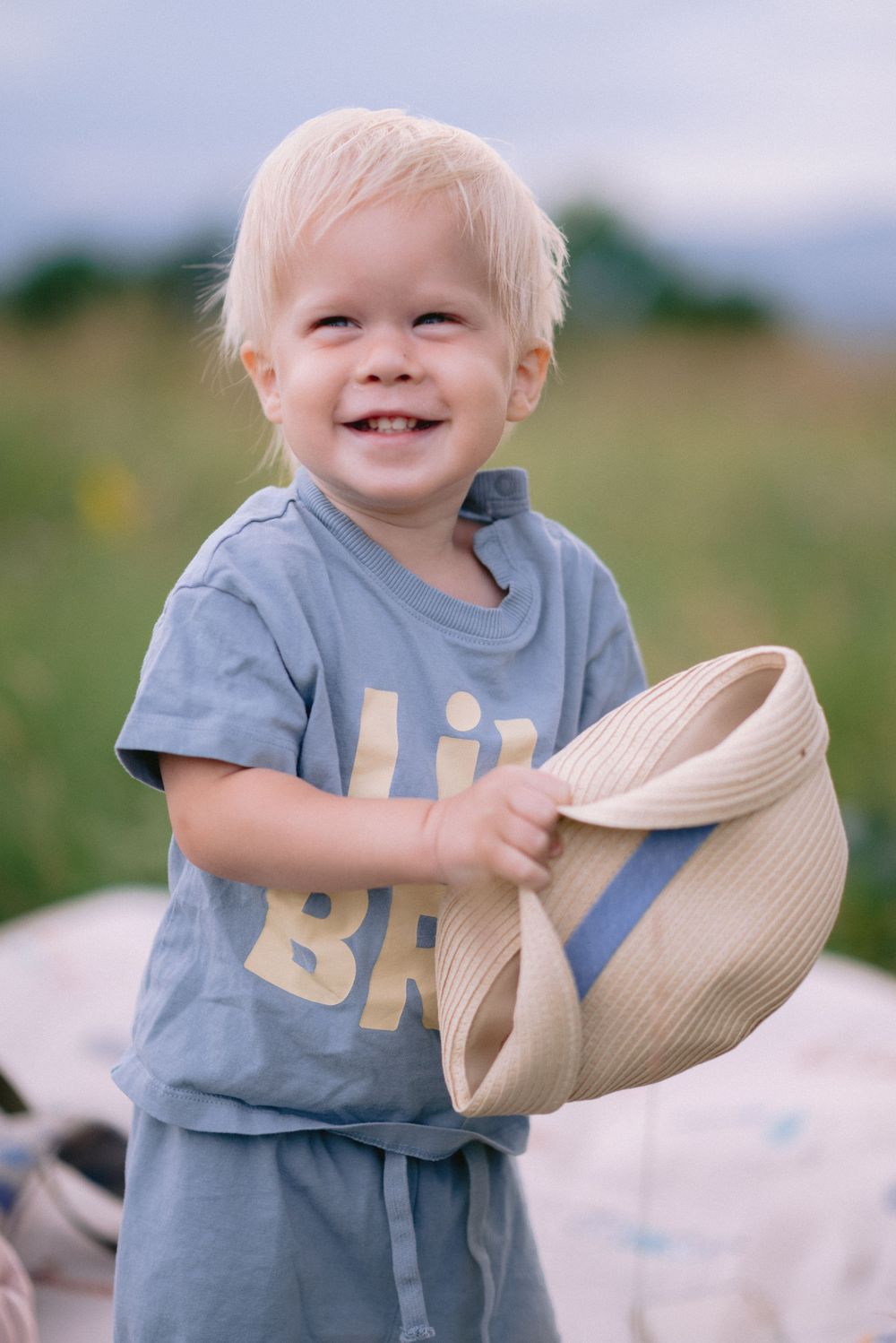 Smiling blonde toddler in a Lil Bro gray shirt holds a straw hat in a sunny outdoor setting.