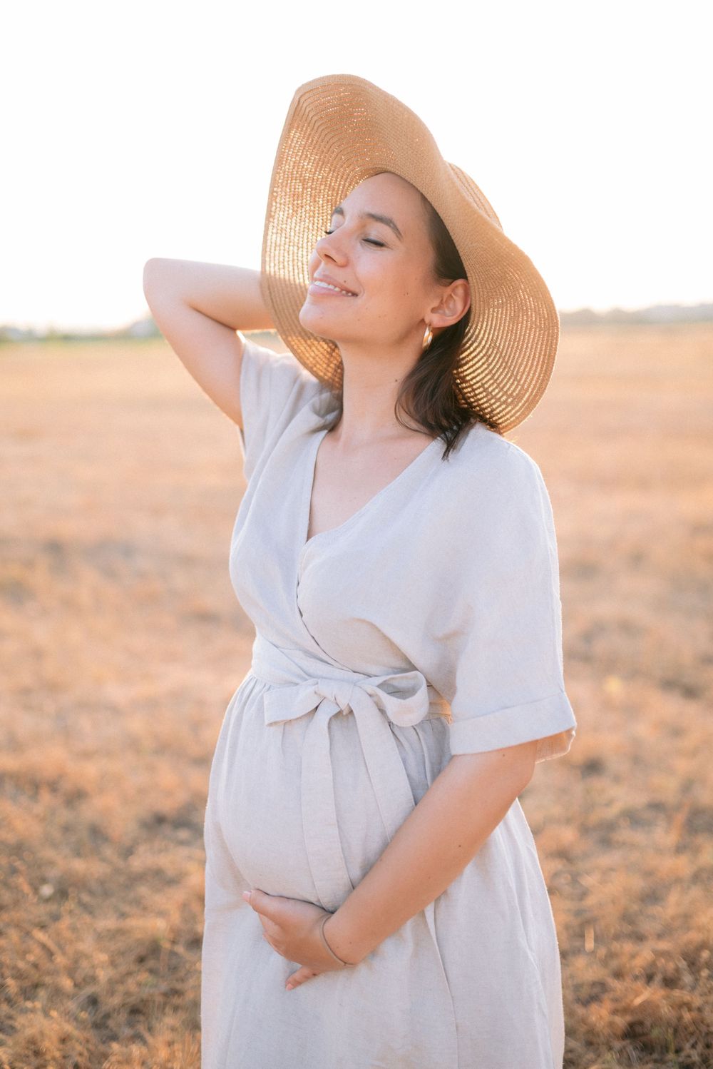 Glowing pregnant woman in linen dress and straw hat cradles her belly in a sunny golden field.
