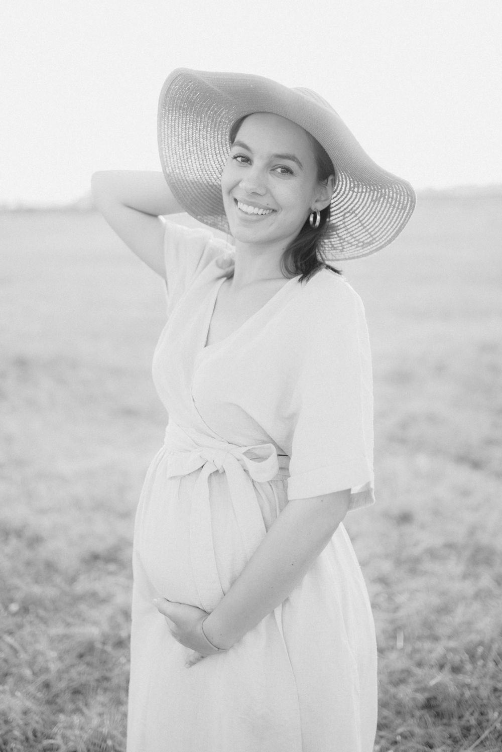 Smiling pregnant woman in white dress and wide-brim hat holds her belly in a field, black and white.