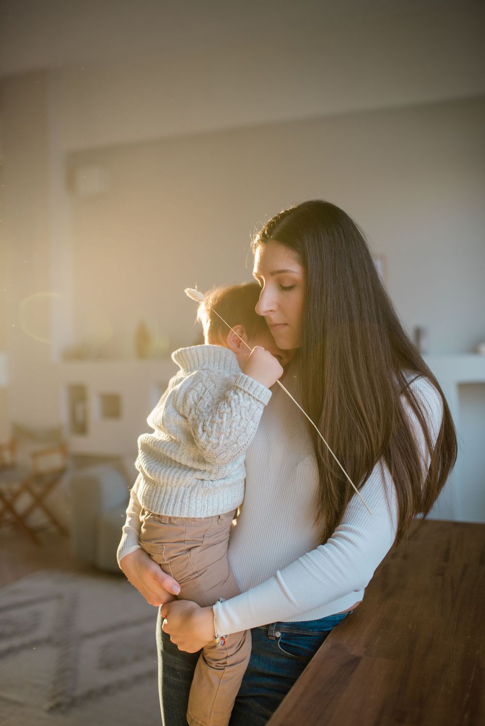 A dark-haired mother tenderly holds her baby in warm golden sunlight indoors.