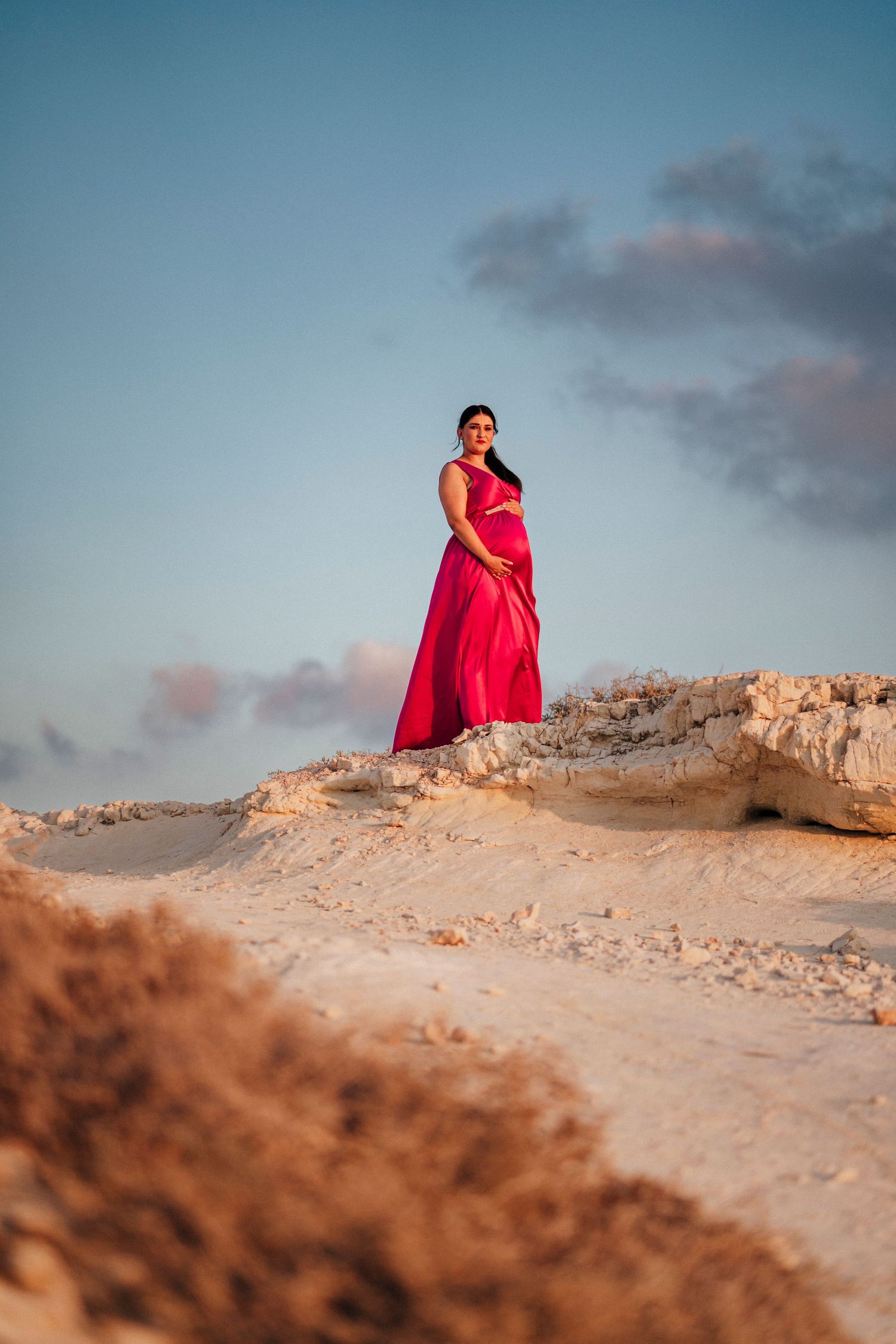 Expectant mother in flowing red dress poses on sandy dunes at sunset.