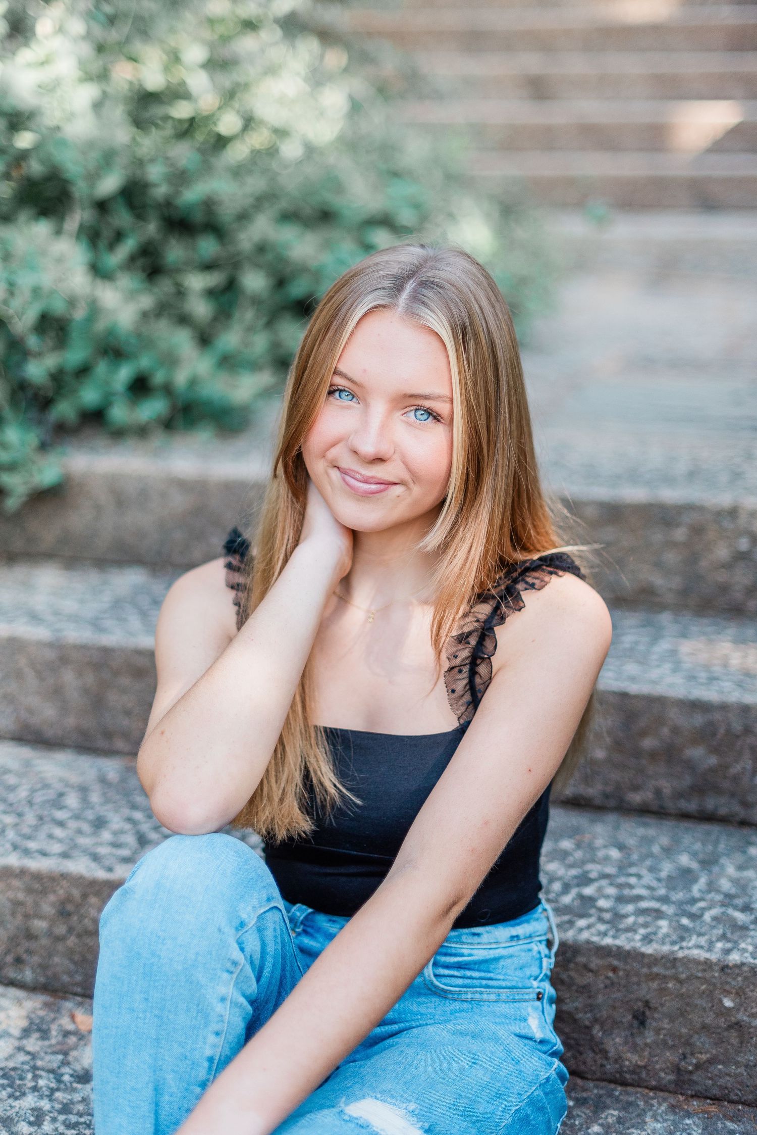 A person in a black tank top and light blue jeans sits casually on outdoor stone steps surrounded by greenery.