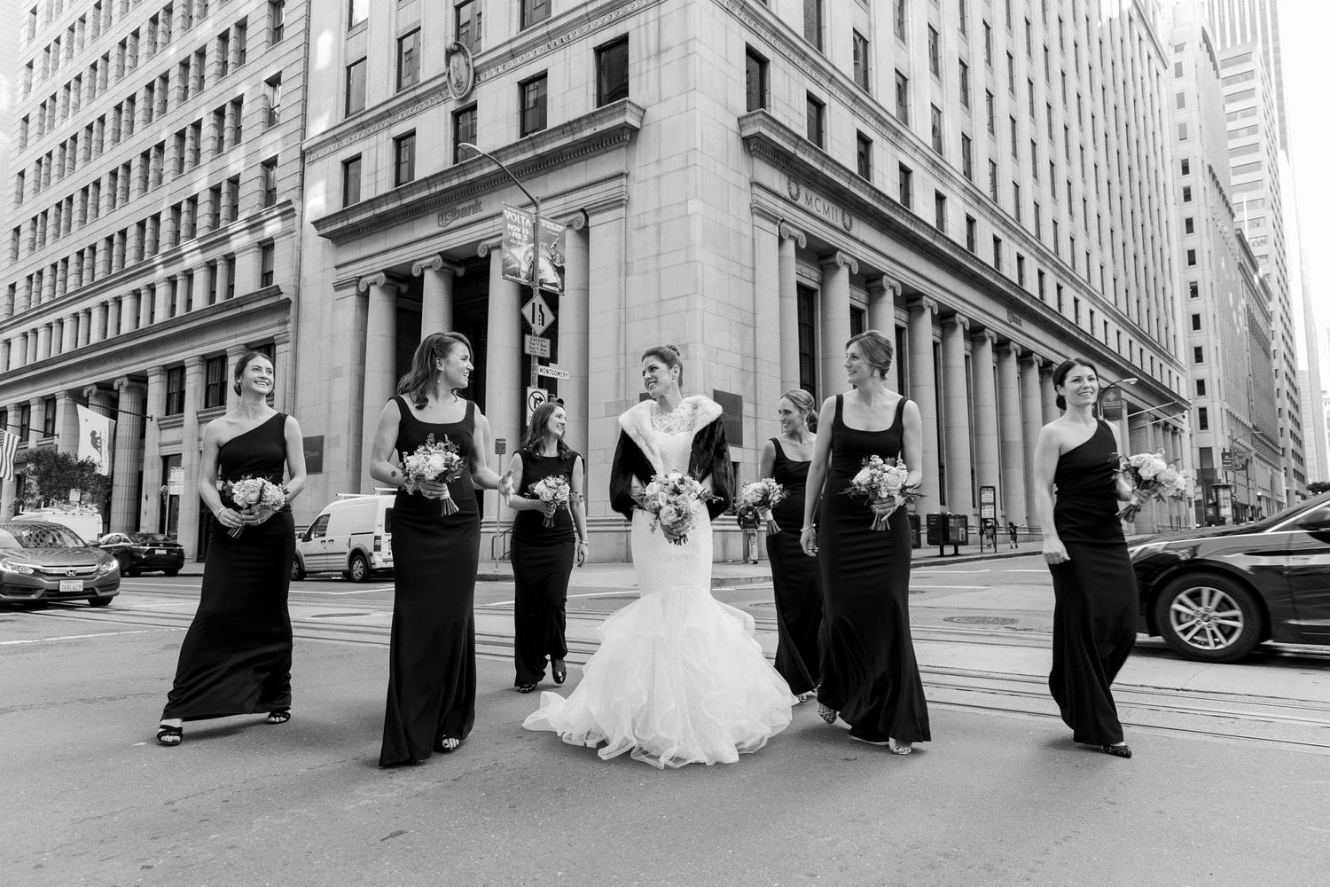 A wedding party poses at an elegant city intersection with bridesmaids in black dresses and a bride in white.