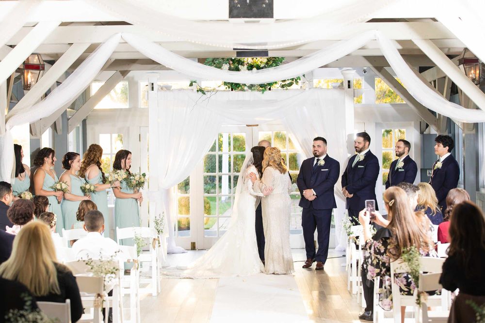 Indoor wedding ceremony in a bright white room decorated with elegant draping and floral accents.