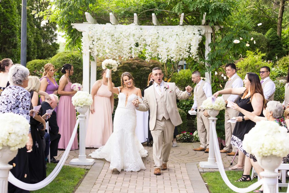 Garden wedding ceremony under white pergola decorated with flowers and white aisle runner.