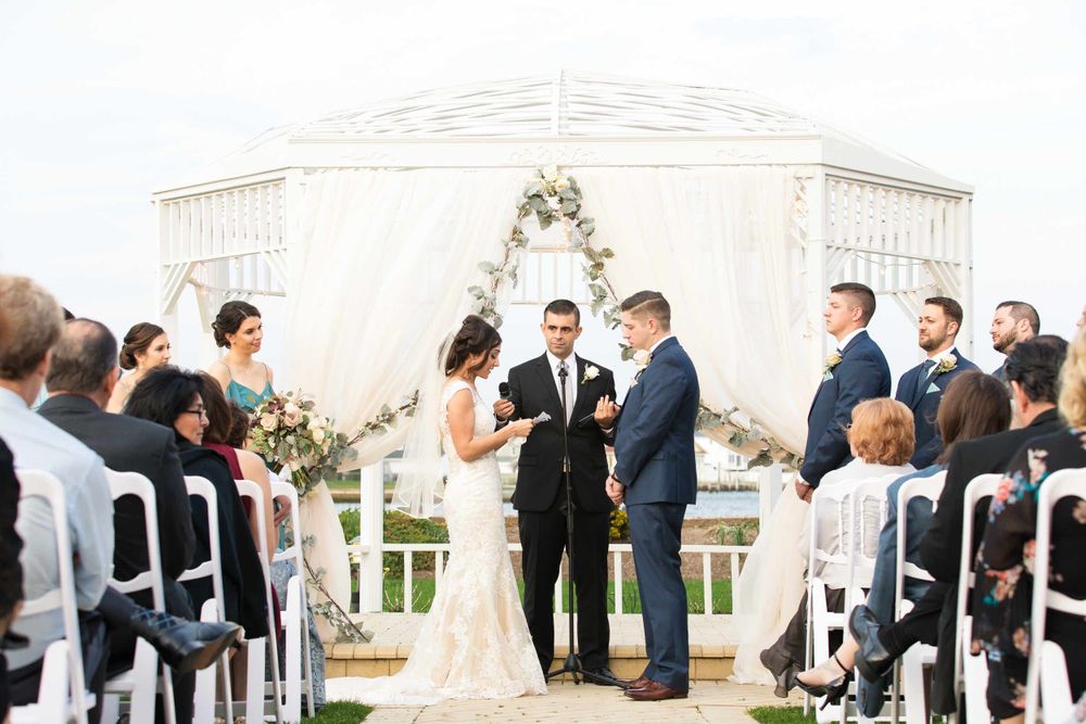 Outdoor wedding ceremony under a white gazebo with floral arch and guests seated on white chairs.