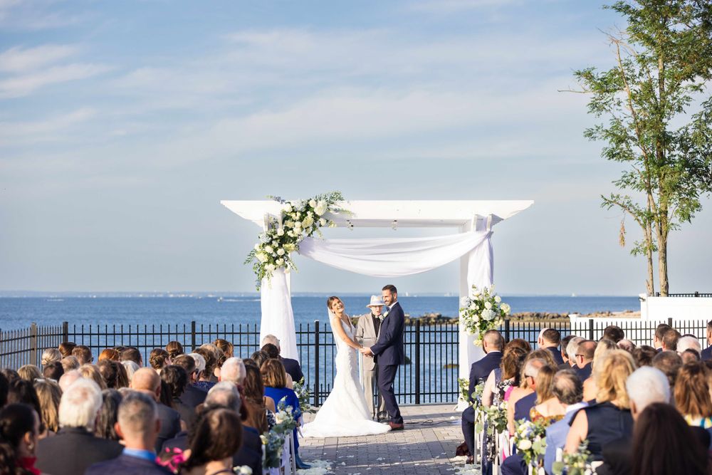 Outdoor wedding ceremony on a waterfront deck with white floral arch and seated guests under blue skies.