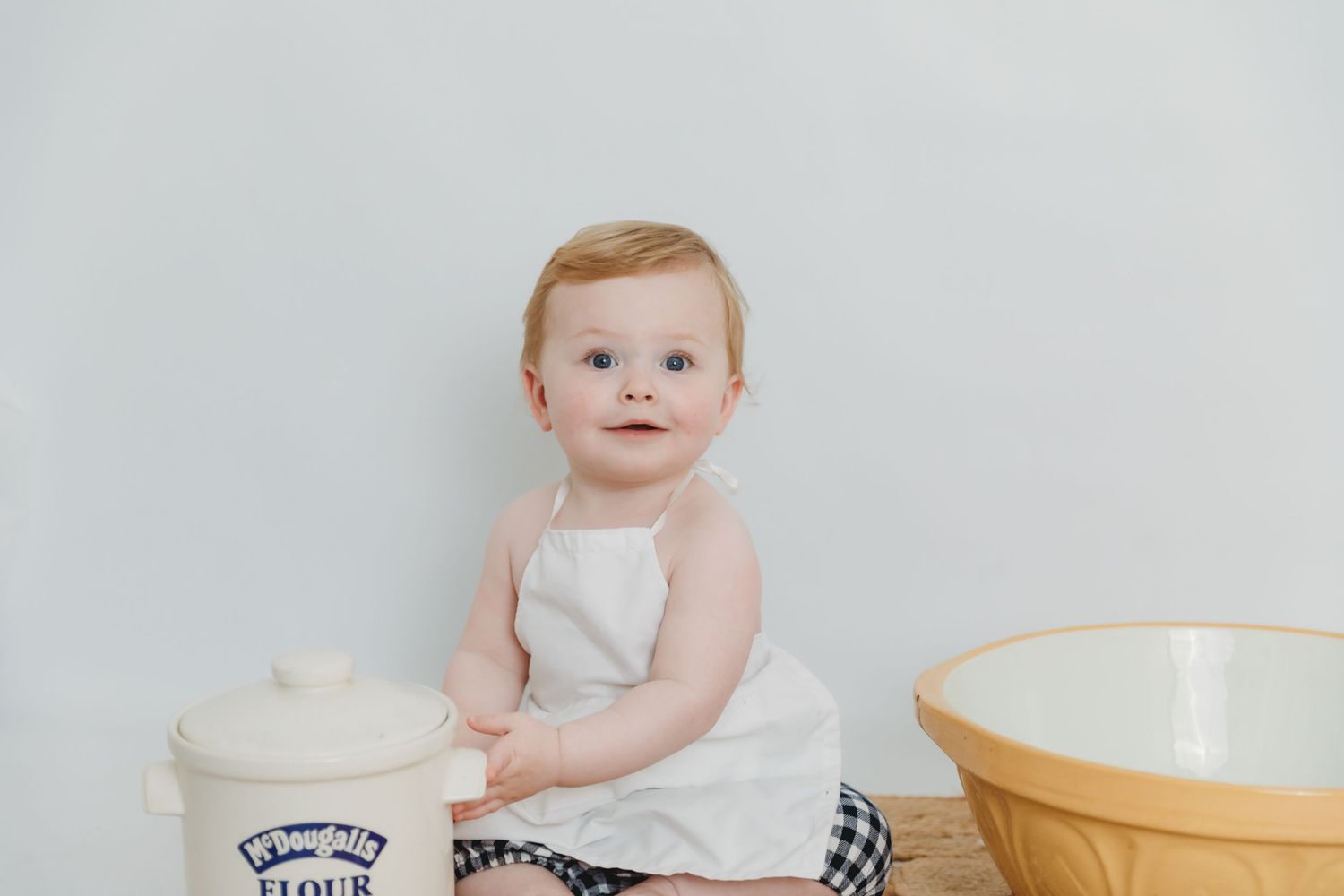 Baby sitting in white onesie next to wooden bowl during milestone photo session.