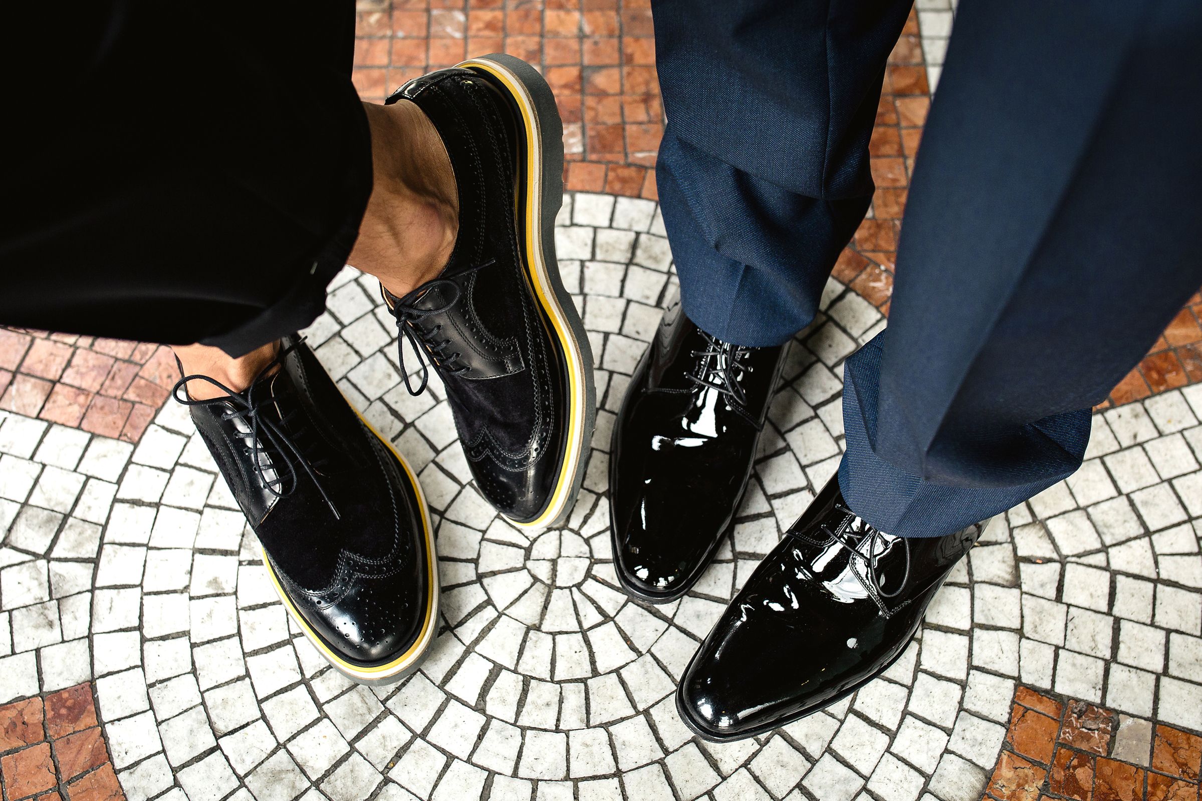High-angle shot of two grooms' feet showing polished black dress shoes on a patterned mosaic floor.