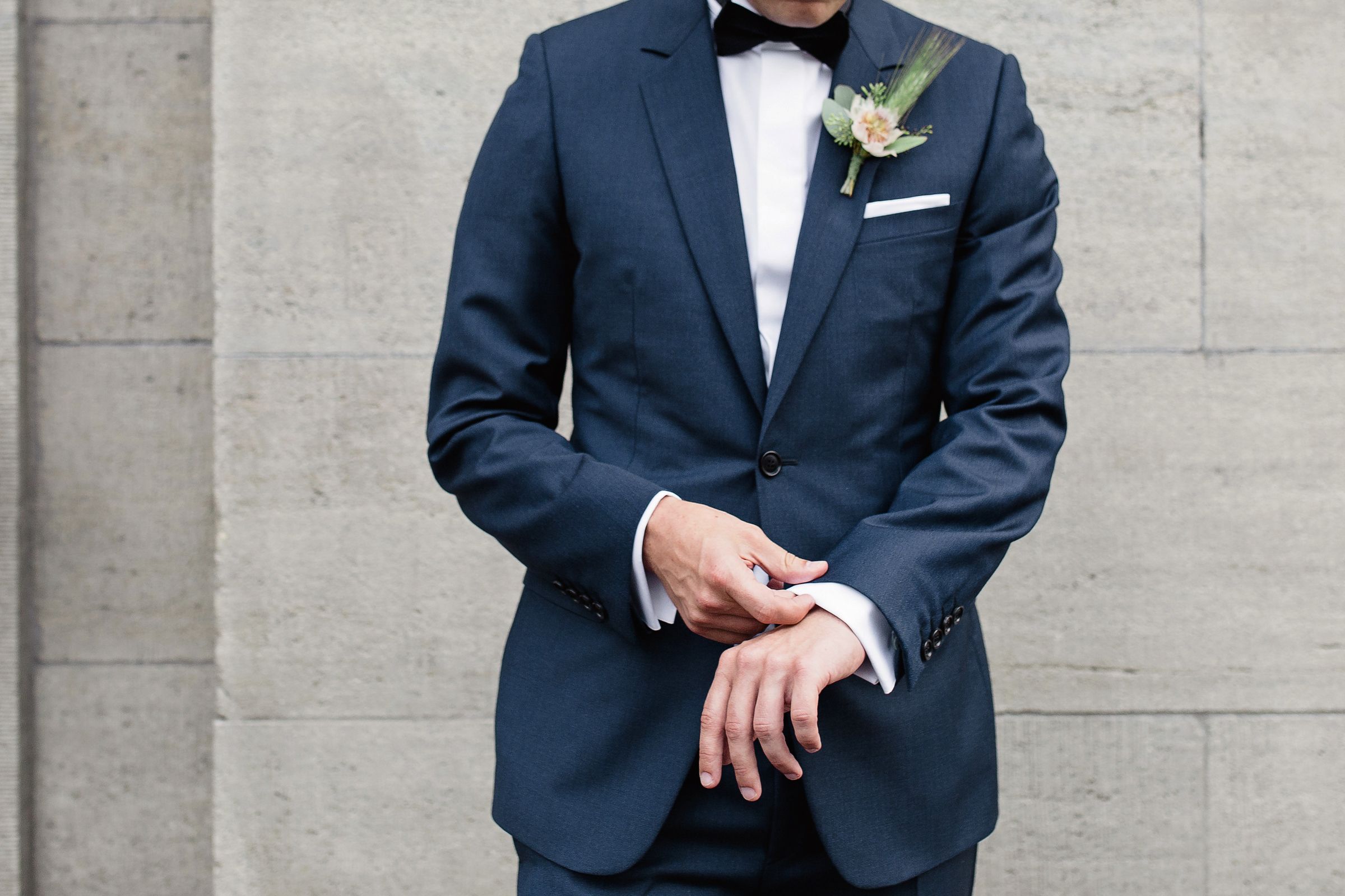 Vertical close-up portrait of a groom in a dark suit, focusing on the floral boutonniere and formal attire.