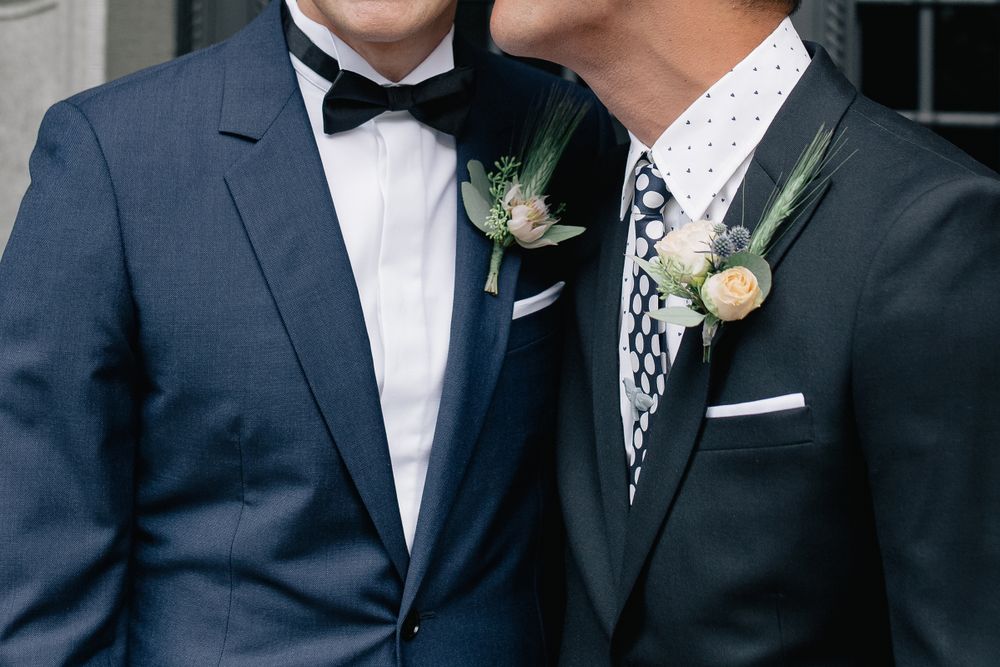 Close-up of two grooms in navy blue suits and bowties with matching floral boutonnieres.