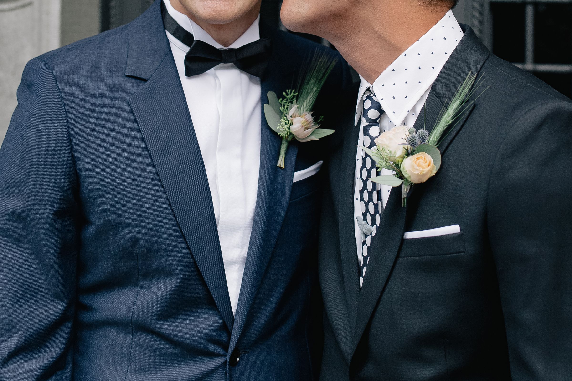 Close-up of two grooms in navy blue suits and bowties with matching floral boutonnieres.