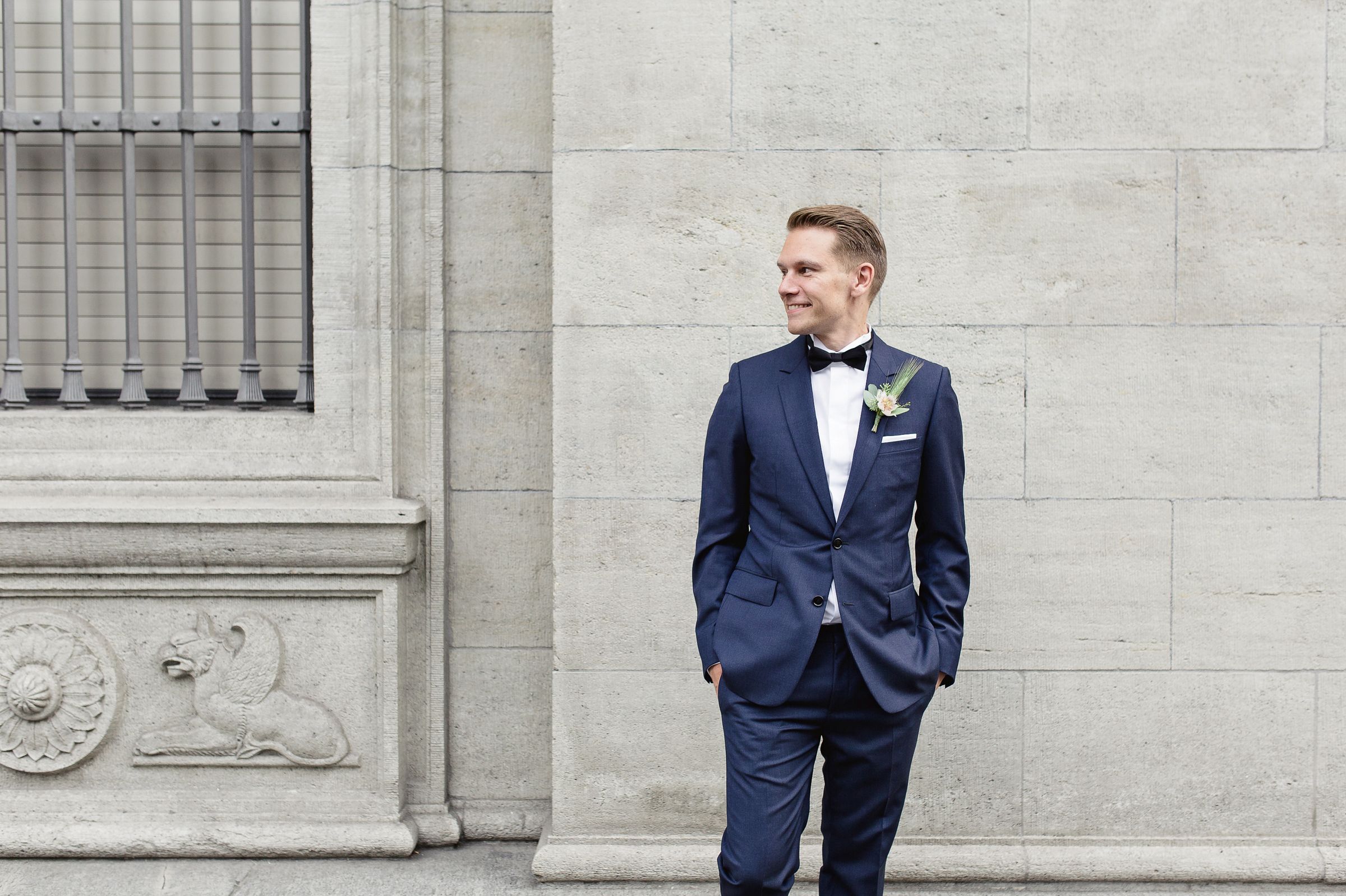 Portrait of a groom in a navy blue suit standing against a classic, light-colored stone wall.