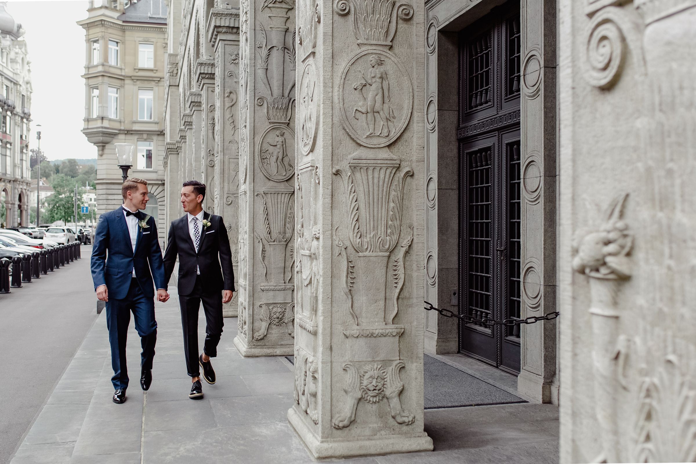 Wide shot of two grooms walking together through a grand stone corridor with historic columns.