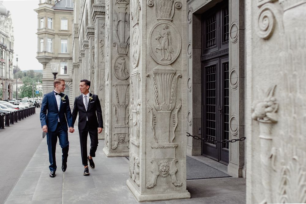 Wide shot of two grooms walking together through a grand stone corridor with historic columns.