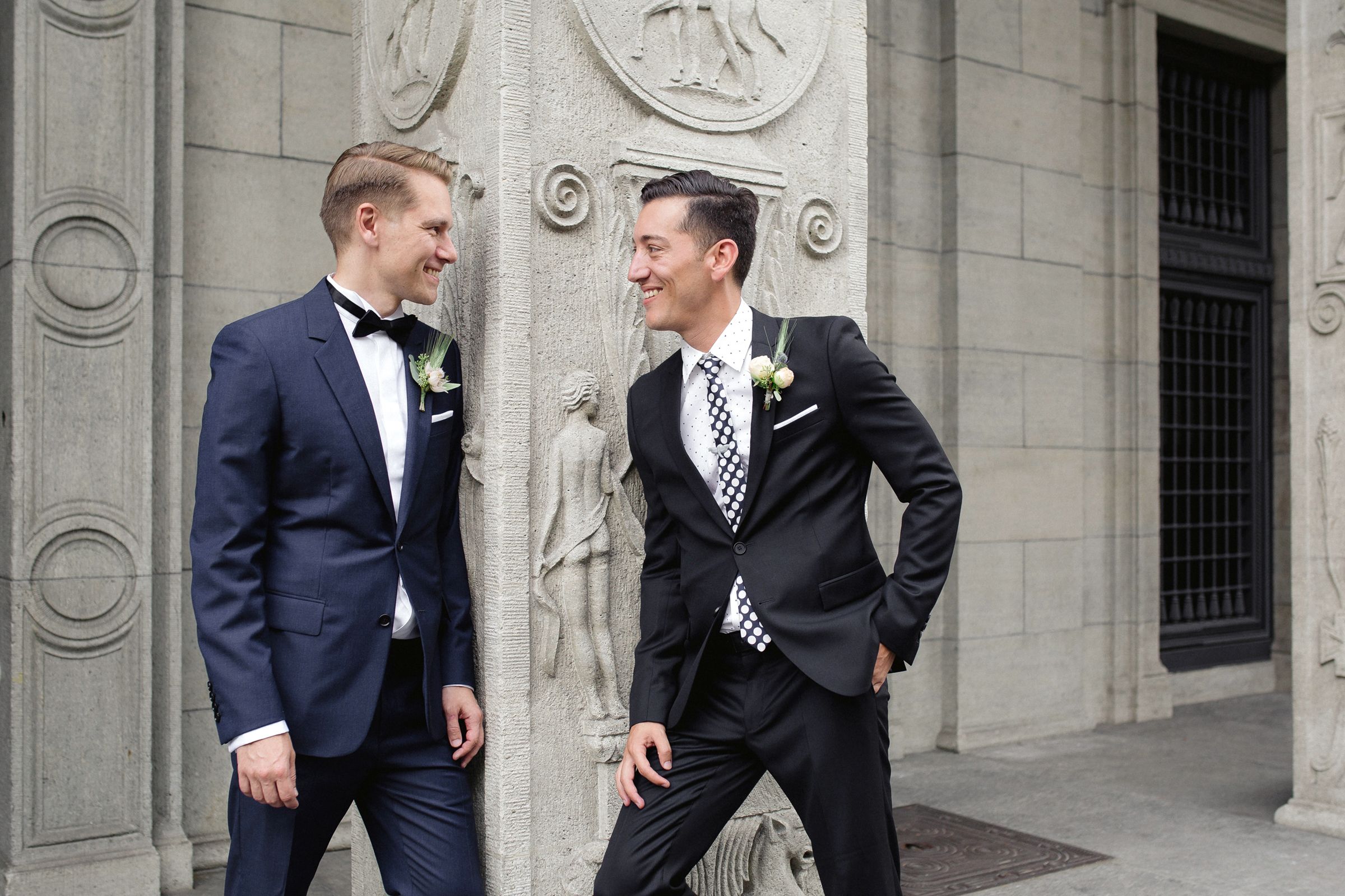 Two grooms in tuxedos sharing a laugh while leaning against a historic stone building.